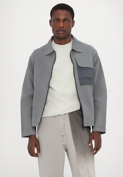 Veste grise avec une fermeture éclair à l'avant, un col montant et une poche poitrine. Portée sur une chemise légère et associée à un pantalon texturé gris clair.
