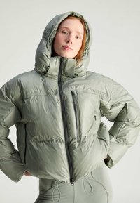 Puffet grøn jakke med hætte, der har en frontlynlås og sidelommer. Materialet virker skinnende med en glat tekstur.