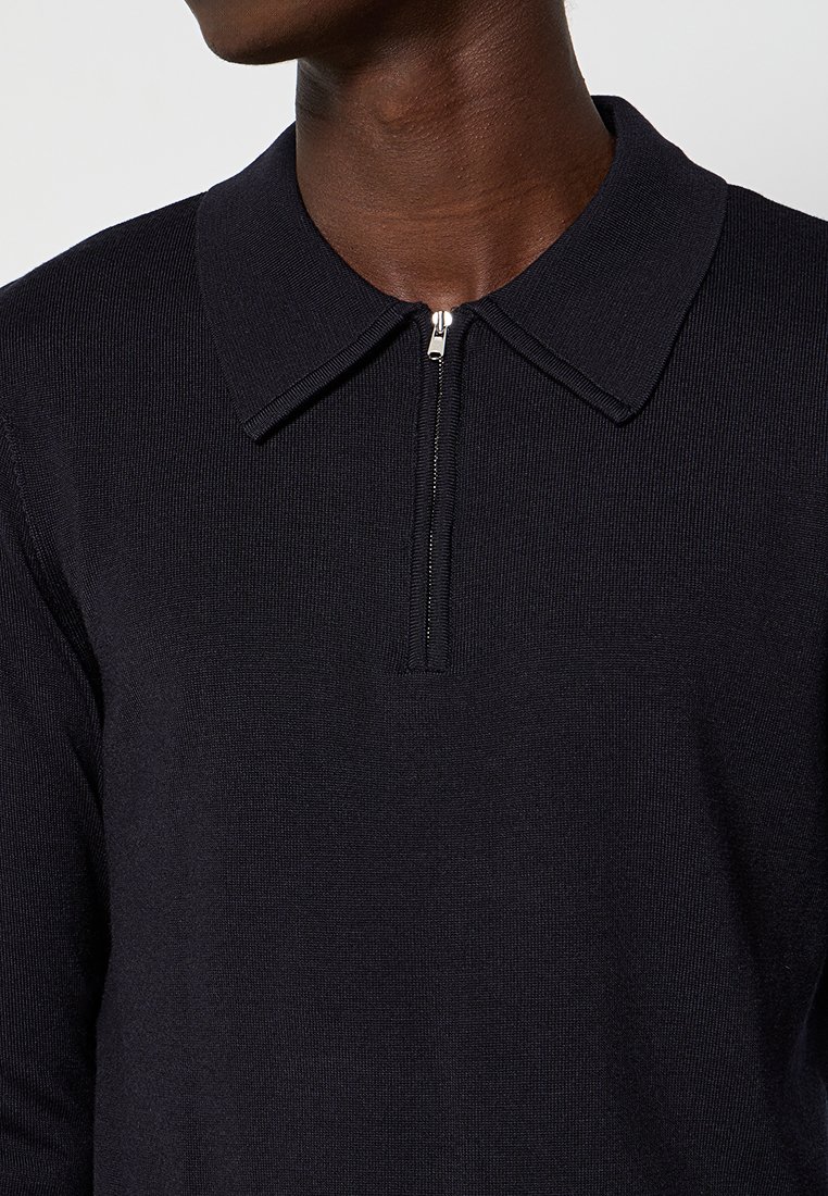Persona de piel oscura que lleva una camiseta de punto de manga larga negra con cuello y una media cremallera en el centro frontal.
