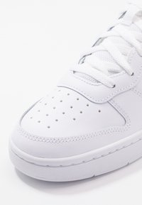 Vit lädersneaker med perforerat tårum, texturerade detaljer, platta snören och en slät gummisula. Minimalistisk design.