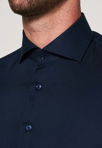 Navy blauwe overhemd met een puntige kraag, voorzien van donkere knopen en een gladde stofstructuur. Close-up weergave benadrukt de stikdetails.