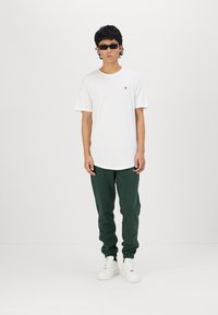 White cotton t-shirt with a small logo, green sweatpants, and white sneakers. The person is wearing black sunglasses and has curly hair.