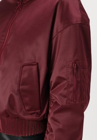 Giacca bomber bordeaux realizzata in tessuto lucido, con chiusura a zip, orlo a coste e numerose tasche, tra cui una con zip.