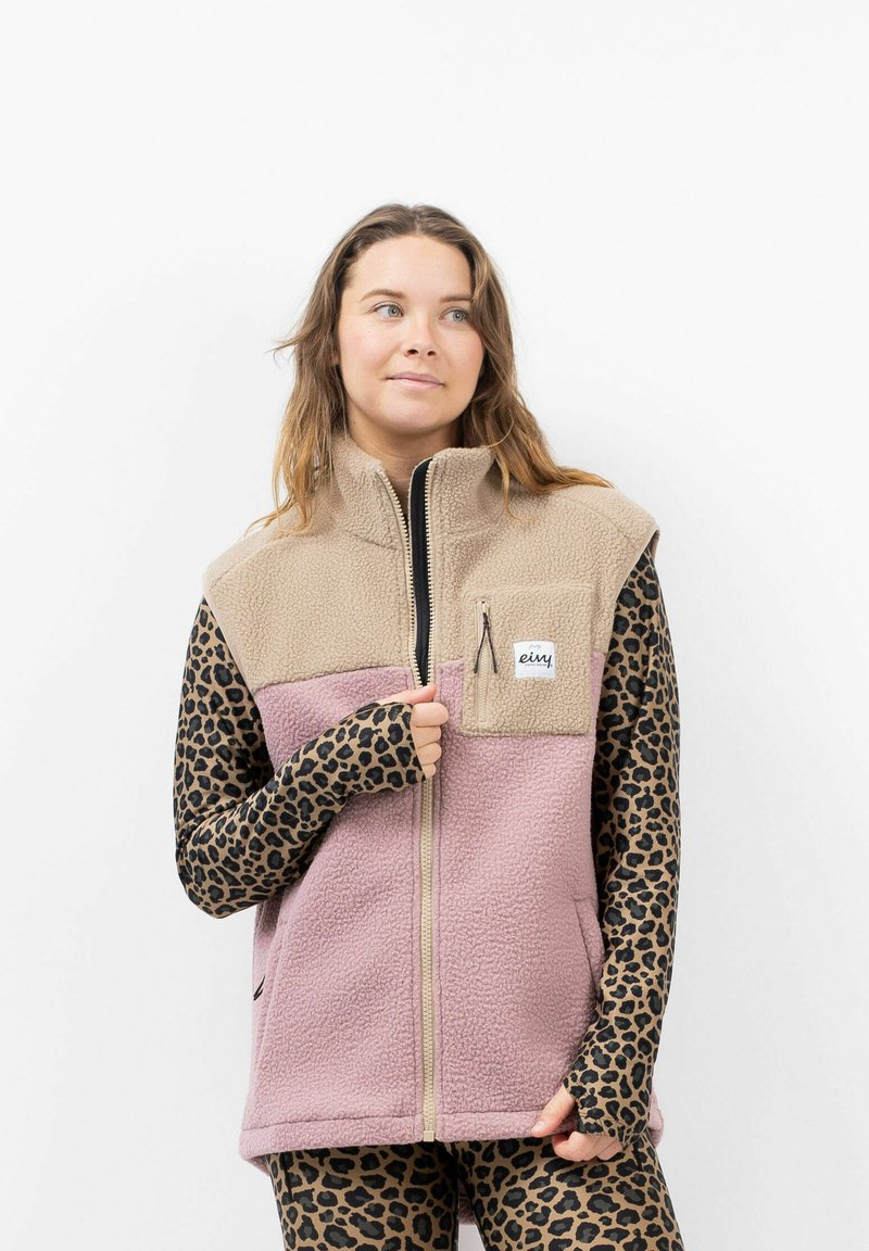 Fleece-Weste mit einem beigen Oberteil und einem pinken unteren Panel, ausgestattet mit einer Reißverschluss-Brusttasche und einem langärmeligen Hemd mit Leopardenmuster darunter.