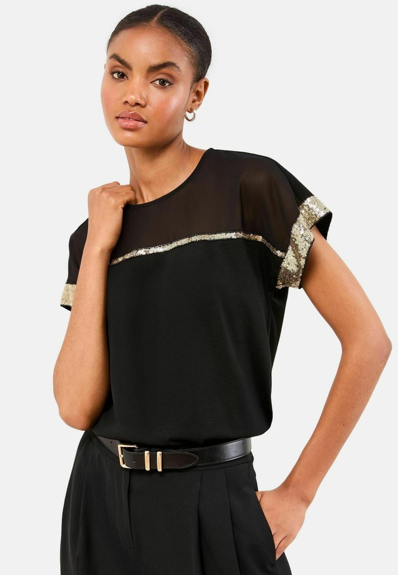 Top negru cu mânecă scurtă, cu un panou transparent negru și accente de paiete aurii pe manșete și piept. Asortat cu pantaloni cu talie înaltă și un curea.
