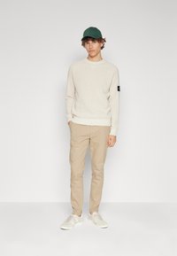 Pull en tricot crème, chinos beige clair et baskets blanches. Casquette verte. Le pull présente une écusson sur la manche gauche. Coupe et style décontractés.