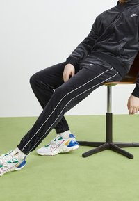 Svart velour-träningsdräkt med vita sidor, zip-up jacka, elastiska ärmslut och matchande byxor. Bärs med flerfärgade sportskor.