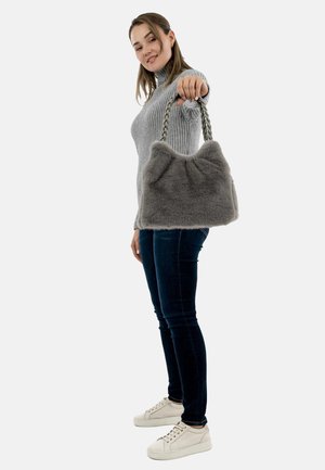 Woman in gray sweater and dark jeans holding out a gray faux fur handbag with braided handles against a white background.
