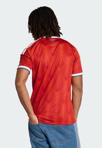 Osoba z dredami ubrana w czerwoną koszulę w wzory na krótkie rękawy z białym i niebieskim obszyciem, z rękami w kieszeniach niebieskich dżinsów, zwrócona tyłem.