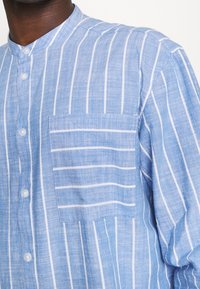 Camicia azzurra chiara con bottoni, realizzata in tessuto testurizzato con righe verticali bianche, dotata di una tasca a toppa sul lato sinistro del petto.