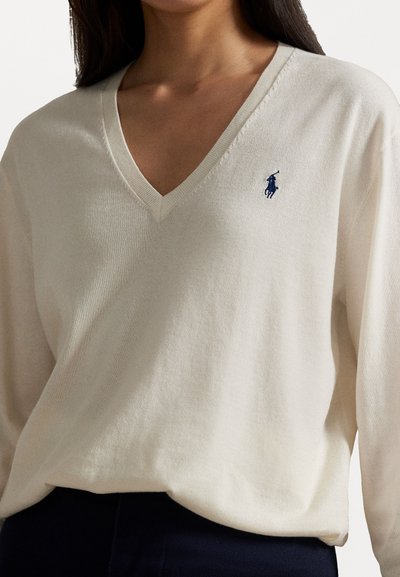 Frau trägt einen cremfarbenen V-Ausschnitt-Pullover mit einem kleinen dunkelblauen Logo eines Polo-Spielers auf der linken Brust, kombiniert mit dunklen Hosen.