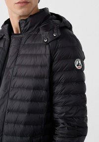 Giacca a vento nera con design trapuntato, frontale con zip e cappuccio rimovibile. Presenta una pettorina rotonda del marchio sulla spalla sinistra.