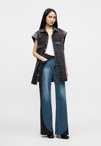 Μαύρο τζιν γιλέκο χωρίς μανίκια με κουμπιά, φορεμένο πάνω από ένα λευκό τοπ, συνδυασμένο με μπλε τζιν παντελόνι φαρδιάς γραμμής με χρωματιστές μπλοκ περιοχές και λεία υφή.