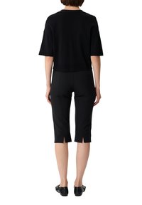 Schwarzes kurzärmliges Oberteil mit geradem Saum, kombiniert mit schwarzen Caprihosen mit seitlichen Schlitzen nahe dem Saum. Flache schwarze Schuhe.