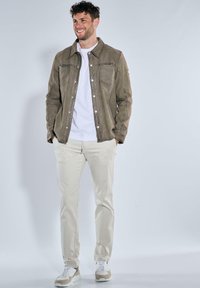 Olivgrüne Wildlederjacke mit klassischem Kragen, zwei Brusttaschen und sichtbaren Nähten, getragen über einem weißen T-Shirt und beigen Hosen.