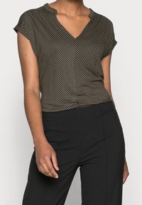 Black and white polka dot blouse with a V-neck, short sleeves, and soft, lightweight fabric, worn with black trousers.