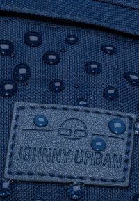 Tengeri kék szövet, melyen kiemelkedő vízálló cseppek találhatóak, egy textúrált sötét szürke bőrfolttal, amelyen "JOHNNY URBAN" felirat és egy logó van bélyegezve.
