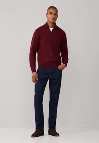 Maglione bordeaux con zip e maniche lunghe, abbinato a jeans blu scuro e scarpe marroni. Il tessuto appare morbido con una superficie liscia.