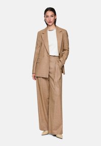 Beige Anzug mit weit geschnittenen Hosen und einem doppelt geknöpften Blazer. Unter dem Blazer ein weißes Oberteil, minimalistisches Design und glatte Textur.