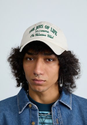 Casquette de baseball beige avec un texte vert foncé indiquant "Les Joies de la Vie, C'est un Mode de Vie, Club de Vacances." Modèle portant une veste en denim.