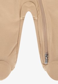 Beige Kleidungsstück mit einem Reißverschluss auf der rechten Seite, mit abgerundeten Kniebereichen, strukturiertem Stoff und glatter Oberfläche.
