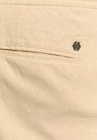Shorts en lin beige avec une texture lisse, arborant un petit logo hexagonal noir sur la poche arrière et un détail de couture subtil.