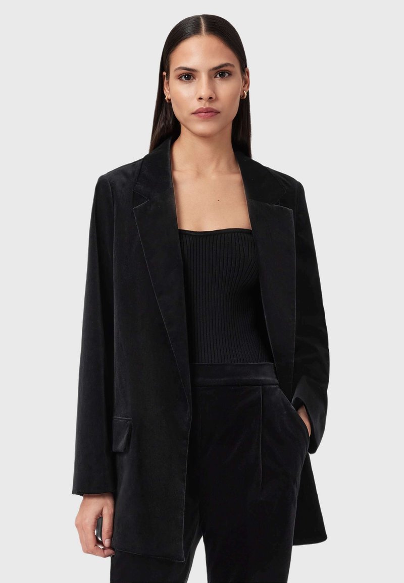 Una donna con capelli scuri e lisci indossa un blazer di velluto nero, un top tubolare a coste nero e pantaloni di velluto nero abbinati, mentre posa contro uno sfondo grigio.