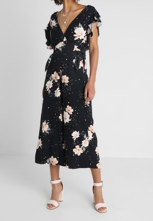 Czarny kombinezon w kwiaty z falbanami na rękawach, z dekoltem w serek, jasnymi wzorami kwiatów i luźnym krojem; zestawiony z różowymi sandałami na paski.