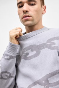 Ung mand med hoops ørepynt og ringe, som holder kraven på en lysegrå sweater med et mørkere grått værktøjsmønster.