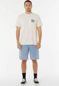 T-shirt beige di cotone con logo blu e arancione sul petto, abbinata a pantaloncini di jeans. Il modello indossa calzini neri e sneakers.