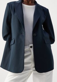 Námořnická modrá sako s jedním knoflíkem, klopami a dvěma předními kapsami, v kombinaci s bílou ribbovanou košilí a krémovými kalhotami.