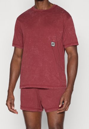 Mann trägt passendes, bordeauxfarbenes, gewaschenes T-Shirt mit kurzen Ärmeln und Shorts mit kleinen schwarz-weißen Logo-Patches auf der linken Seite.