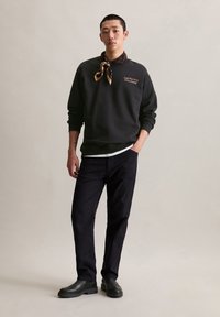 Felpa nera con logo, indossata sopra una camicia bianca. Abbinata a pantaloni scuri e stivali neri. Sciarpa con motivo attorno al collo.