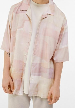 Persona che indossa una camicia a maniche corte, abbottonata, rosa pastello e beige con motivo sopra una T-shirt bianca, con pantaloni chiari.