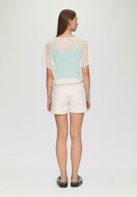 Witte mesh top met korte mouwen over een lichtblauw hemd, gecombineerd met beige opgerolde shorts en zwarte instapsandalen.