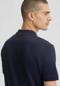 Camisa polo de color azul marino con cuello que presenta una línea de pequeños adornos; mangas cortas y textura suave y lisa.