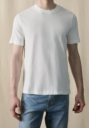 Mężczyzna ubrany w gładką białą koszulkę z krótkim rękawem i okrągłym dekoltem, włożoną do jasnoniebieskich jeansów, na neutralnym tle.