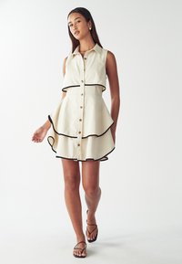 Giovane donna che indossa un abito senza maniche color crema con bordi neri, con bottoni frontali e una gonna a strati e svasata, che cammina con sandali neri con cinturini su sfondo bianco.