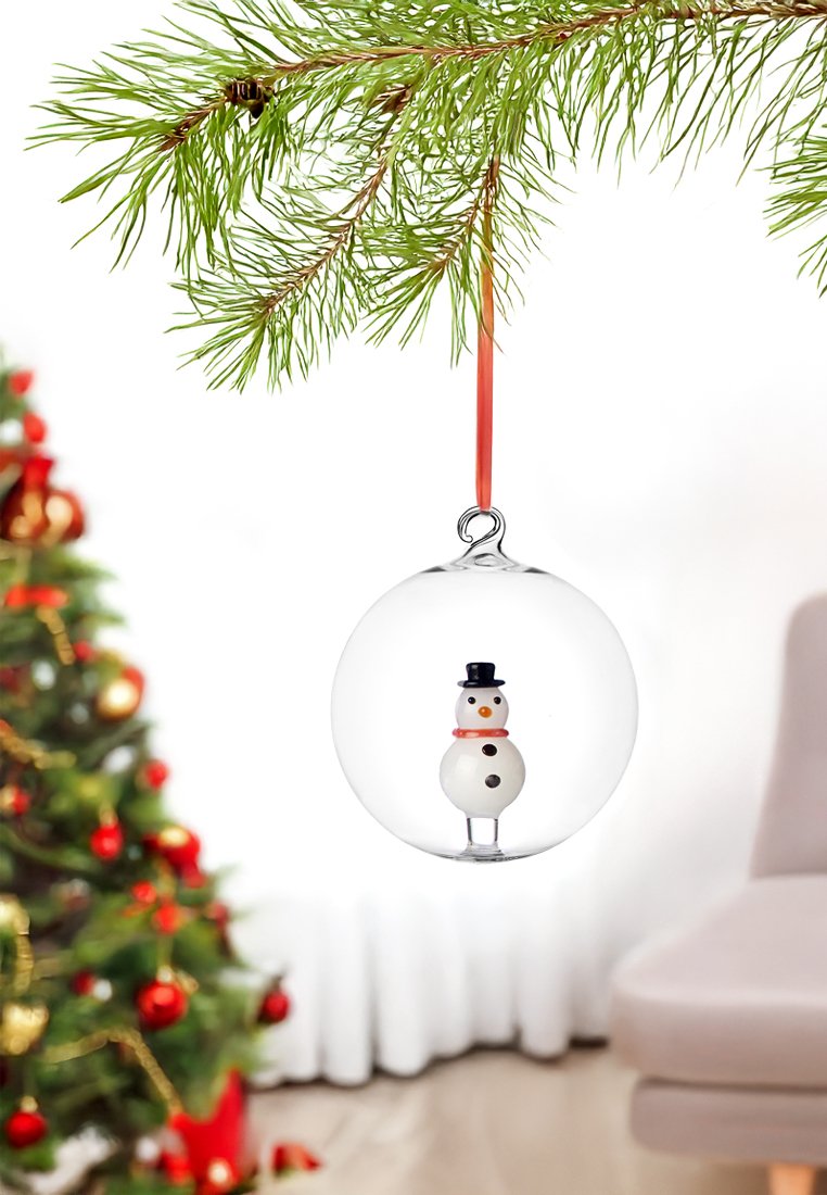 Durchsichtige Weihnachtsornament aus Glas mit einem weißen Schneemann, der einen schwarzen Hut und einen orangefarbenen Schal trägt, der an einem roten Band von einem Tannenzweig hängt.