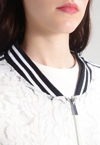 Bílá bomber bunda s krajkovou vrstvou, pruhovaný černobílý žebrovaný límec, zipové zapínání a roztrhané okraje, nošená přes světlo barevnou košili.