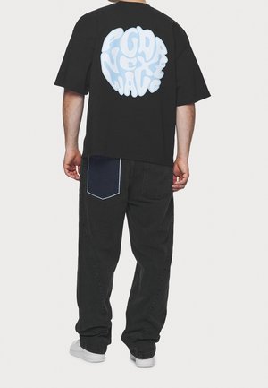 Camiseta negra de gran tamaño con texto ondulado de color azul claro en la espalda, combinada con vaqueros negros que tienen un bolsillo de parche de mezclilla y zapatillas blancas.