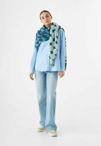 Lichtblauw overhemdshemd, brede jeans en een grote geklede sjaal met cirkels en driehoeken in blauw en groene tinten.