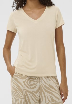 Vrouw draagt een effen beige V-hals T-shirt met korte mouwen en beige broek met een lichtbruin zebraprintpatroon, staand met één hand in de zak.