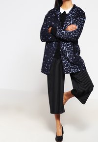 Donna in piedi su una gamba e a piedi nudi, indossa un cappotto con stampa leopardata blu, un maglione nero, una camicia con colletto bianco e pantaloni larghi a righe nere.