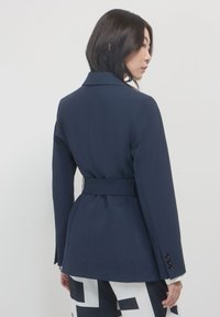 Donna con capelli neri fino alle spalle, che indossa un blazer blu marina con cintura e pantaloni a righe in bianco e nero, girata di lato su uno sfondo neutro.