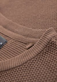 Suéter marrón de punto que presenta un patrón texturizado, cuello acanalado y una pequeña etiqueta. El material tiene una apariencia suave y ligeramente burbujeante.
