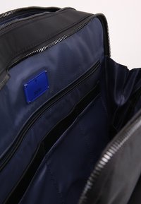 Svart tygryggsäck med blå foder, utrustad med flera zippered fickor och en märkt blå etikett. Slät textur, modern design.