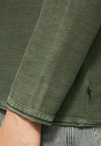 Haut à manches longues vert olive en tissu texturé. Doté de poignets et d'un ourlet côtelés, d'une étiquette sur la couture et d'une coupe décontractée.