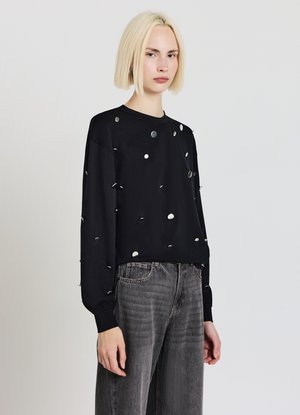 Schwarzes Langarmshirt mit runden weißen und silbernen Akzenten, aus weichem Stoff. Kombiniert mit grauen, weit geschnittenen Jeans für einen lässigen Look.