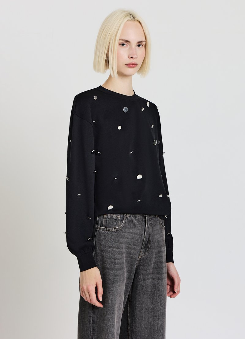 Schwarzes Langarmshirt mit runden weißen und silbernen Akzenten, aus weichem Stoff. Kombiniert mit grauen, weit geschnittenen Jeans für einen lässigen Look.